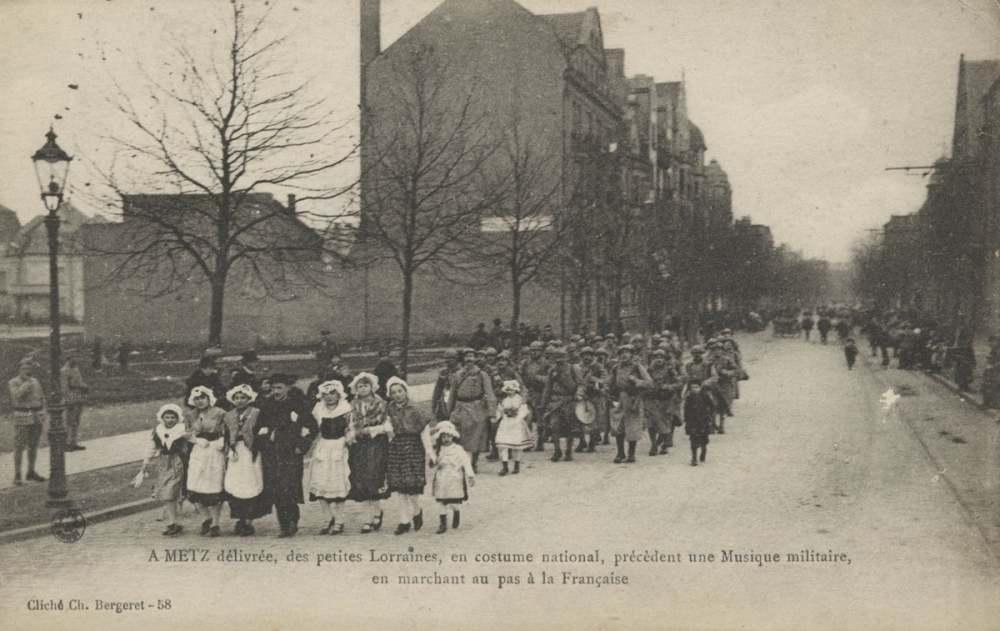 A Metz délivrée, des petites Lorraines, en costume national, précèdent une Musique militaire, en marchant au pas à la Française.