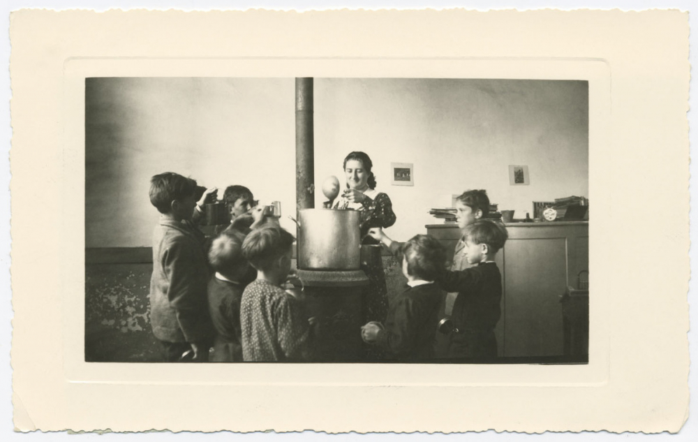 Voilà l'heure de la soupe... Et la souriante institutrice distribue aux petits paysans affamés le bouillon qui, pendant la classe du matin, a tout simplement cuit sur le poële. Pas de matériel, pas de frais, mais des résultats avec un peu de dévouement et de bonne humeur.