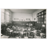Ecole maternelle. La classe des grands : tables individuelles avec tiroirs ; disposition en demi-cercle. Des animaux, des fleurs, des plantes et le ménage complet de la poupée. 