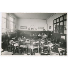 Ecole maternelle. La classe des grands : tables individuelles avec tiroirs ; disposition en demi-cercle. Des animaux, des fleurs, des plantes et le ménage complet de la poupée.