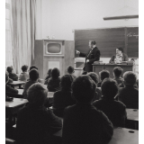 Enseignement audio-visuel. Une classe et deux adultes regardent un poste de télévision installé dans un coin de la pièce, près du tableau noir. 