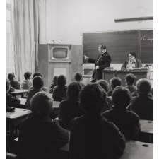 Enseignement audio-visuel. Une classe et deux adultes regardent un poste de télévision installé dans un coin de la pièce, près du tableau noir.