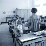 Ecole primaire. L'institutrice, en blouse, vérifie les ardoises.