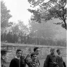 Rentrée scolaire, 1956 : élèves, le cartable sous le bras, dans la cour de l'école ; les parents observent depuis la rue