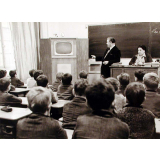 Enseignement audio-visuel. Une classe, deux adultes regardent un poste de télévision installé dans un coin de la pièce, près du tableau noir