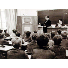 Enseignement audio-visuel. Une classe, deux adultes regardent un poste de télévision installé dans un coin de la pièce, près du tableau noir