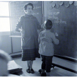 Ecole primaire. L'institutrice aide un élève à écrire au tableau.
