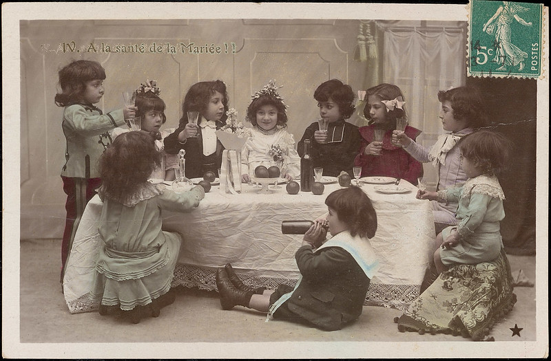 Photo noir et blanc montrant 10 enfants, âgés de 3 ans environ, autour d'une table avec une nappe, en tenue de cérémonie de mariage. Tous ont un verre à pied à la main.  Un enfant est assis de profil devant la nappe ; il boit à la bouteille. 
