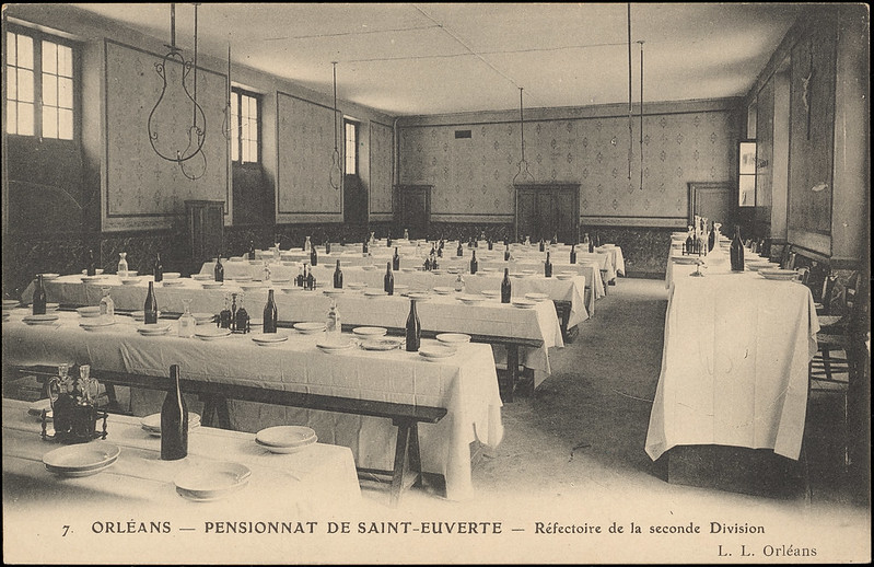 Salle de réfectoire dans laquelle les tables sont dressées avec nappes blanches, vaisselle, carafe d'eau et bouteille d'alcool. 