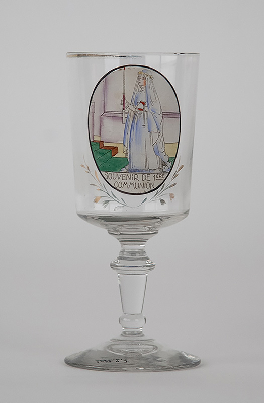 Verre à pied décoré d'un dessin coloré représentant une jeune fille en tenue religieuse tenant un cierge allumé dans sa main droite et un livre et un chapelet dans sa main gauche. elle est devant deux marches. Sous le dessin, il est écrit "Souvenir de première communion".