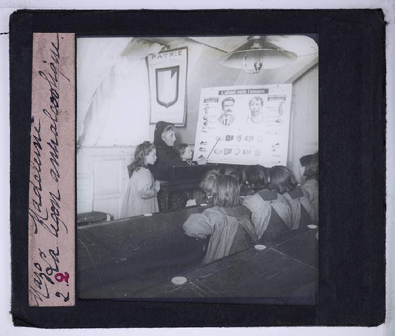 Classe de filles assistant à une leçon antialcoolique. L'enseignante montre avec une baguette une planche didactique intitulée "L'alcool, voilà l'ennemi".