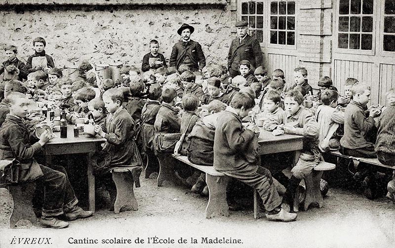 Il s'agit d'une photo en noir et blanc datant de 1905 montrant 3 grandes tablées de garçons en train de déjeuner en extérieur. Ils sont sûrement dans l'enceinte de l'établissement scolaire. On voit des bouteilles d'alcool sur les tables. Certains élèves regardent le photographe.